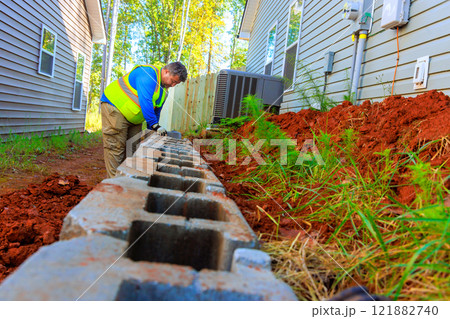 Worker focuses on placing retaining wall blocks next to house in quiet suburb with lush greenery. 121882740