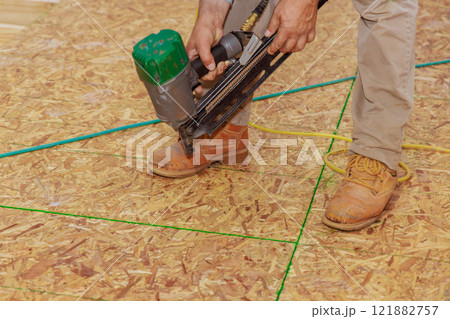 Carpenter worker operates air nail gun to fasten boards as part of flooring installation in construction project. 121882757