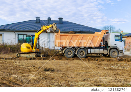 Excavator operates at construction site, with an excavator loading dirt into dump truck beside house. 121882764