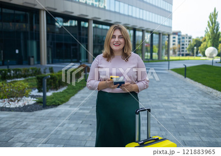 Smiling Caucasian 30 years old business woman near business center using smartphone With travel suitcase, using smartphone, waiting for transport, standing outdoors 121883965