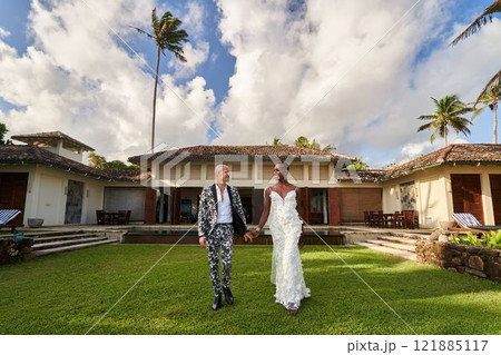 Gender fluid person in bridal dress, LGBTQ groom in bold suit at tropical wedding villa. Happy couple walks hand in hand on grass, celebrating love, diversity, inclusion at unique outdoor venue. Gender fluid person in bridal dress, LGBTQ groom in bold suit at tropical wedding villa. Happy couple walks hand in hand on grass, celebrating love, diversity, inclusion at unique outdoor venue. 121885117