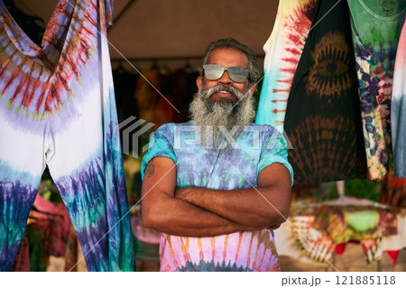 Senior man with long beard wearing tie-dye t-shirt at outdoor craft fair. Smiling, arms crossed, surrounded by colorful clothes. Elderly artist shares young spirit, creative lifestyle. Bohemian event. 121885118