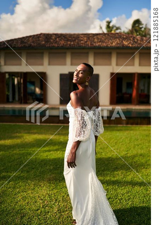 Smiling person in white wedding dress walks on villa lawn under blue sky. Gender fluid black bride celebrates LGBTQ wedding. Summer outdoor ceremony. Elegant bridal fashion. 121885168