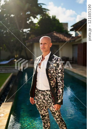Bald man in bold floral suit stands at villa poolside. Daytime event, stylish attire with open shirt. Fashion trend. Possible gay groom outfit. Elegant garden setting. Bald man in bold floral suit stands at villa poolside. Daytime event, stylish attire with open shirt. Fashion trend. Possible gay groom outfit. Elegant garden setting. 121885190