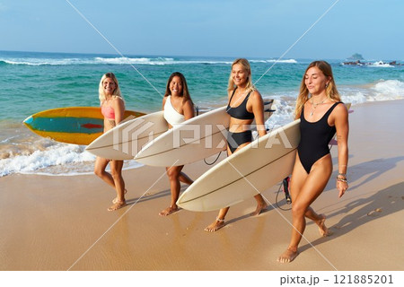 Four women carry surfboards along sandy beach. Girls in swimsuits, smiling, walk by ocean water. Friends ready for surfing adventure at tropical seaside. Warm sunny day, blue sky. 121885201