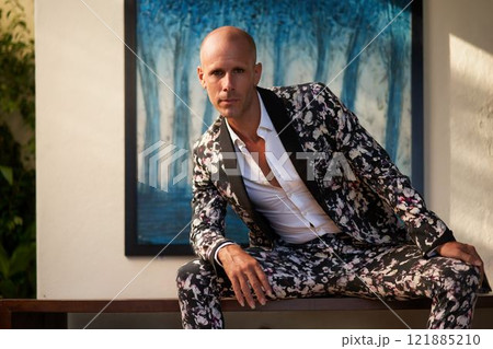 Bald man in floral suit sits confidently at luxury villa. Bold fashion wedding style, groom attire concept. Sunlit elegant setting. Sophisticated outfit trend for modern nuptials. Bald man in floral suit sits confidently at luxury villa. Bold fashion wedding style, groom attire concept. Sunlit elegant setting. Sophisticated outfit trend for modern nuptials. 121885210
