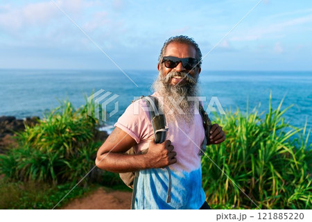 Smiling elderly man with backpack stands near ocean view. Bearded traveler in sunglasses enjoys nature adventure on sunny day. Senior male exploring coastal trail. Outdoor travel lifestyle. Smiling elderly man with backpack stands near ocean view. Bearded traveler in sunglasses enjoys nature adventure on sunny day. Senior male exploring coastal trail. Outdoor travel lifestyle. 121885220