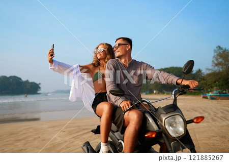 Couple on tropical beach ride scooter. Woman in bikini, guy in casual shirt. Take selfie, smile on sandy ocean shore. Travel influencers explore exotic place. Adventure, freedom mood. 121885267