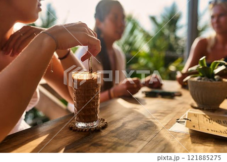 Group of friends relax in outdoor cafe with iced coffee drinks. Sunlit patio with rich plants. People chat, enjoy beverages. Casual meeting or social gathering. 121885275