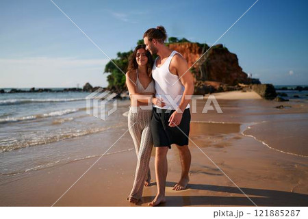 Couple strolls on sandy beach at sunset, man embracing woman, romantic walk by the sea. Pair explore tropical coast, bonding, casual summer clothes. Love leisure lifestyle holiday concept. Couple strolls on sandy beach at sunset, man embracing woman, romantic walk by the sea. Pair explore tropical coast, bonding, casual summer clothes. Love leisure lifestyle holiday concept. 121885287