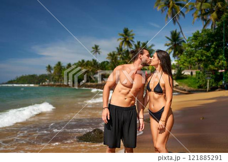 Romantic couple kissing on tropical beach, man and woman in swimwear enjoying honeymoon by sea. Fit pair with toned bodies sharing tender moment under palm trees, love on sunny coastline. 121885291