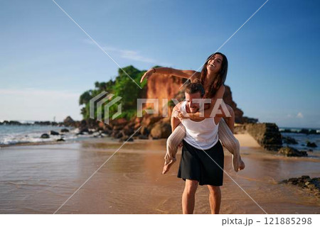 Joyful woman gets piggyback ride from man on beach. Couple enjoys tropical getaway, sandy shore near sea. Playful partner points in distance. Romantic coastal vacation, fun in sun, love moments. 121885298