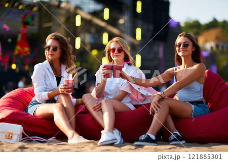 Three young women sit on red bean bag chairs, toast with colorful drinks at a beachside music festival. Friends enjoy a sunny social gathering, casual fashion, fun atmosphere, live event background. Three young women sit on red bean bag chairs, toast with colorful drinks at a beachside music festival. Friends enjoy a sunny social gathering, casual fashion, fun atmosphere, live event background. 121885301