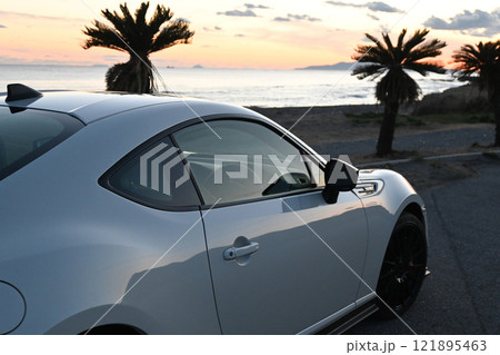 夕焼けの海沿いに止まる、白いスポーツカー 121895463