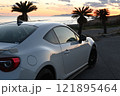 夕焼けの海沿いに止まる、白いスポーツカー 121895464