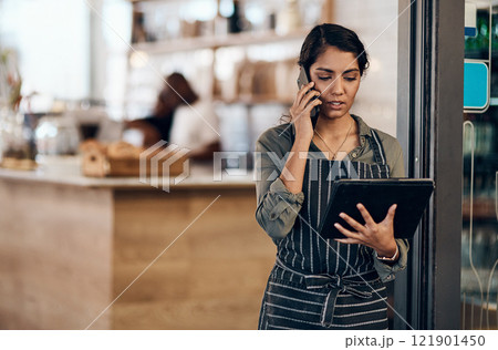 Female cafe business owner using phone talking, ordering and reading on tablet in her store. Serious businesswoman, entrepreneur or employee standing in a coffee shop preparing online grocery order. 121901450