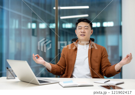 Mature Asian man resting meditating sitting in lotus position inside office. Businessman working with laptop at workplace, doing breathing exercises. 121902417