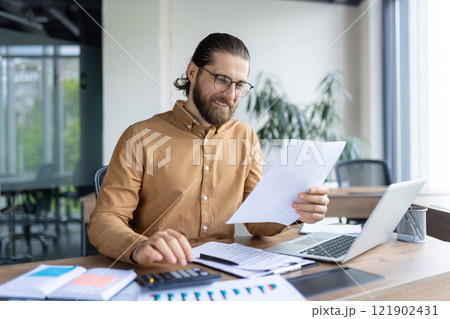 A focused professional reviews paperwork while working on financial or business tasks at a desk with a laptop and calculator, suggesting determination, productivity, and a modern office environment. A focused professional reviews paperwork while working on financial or business tasks at a desk with a laptop and calculator, suggesting determination, productivity, and a modern office environment. 121902431