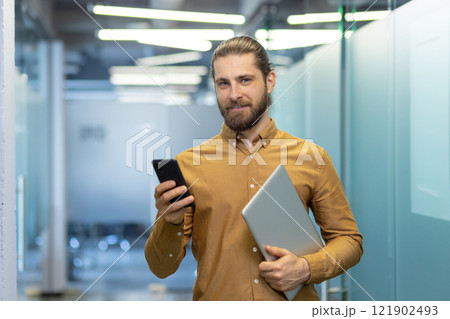Young professional dressed in business casual attire holds a laptop and smartphone while standing in a modern office environment, showcasing confidence and tech-savviness. 121902493