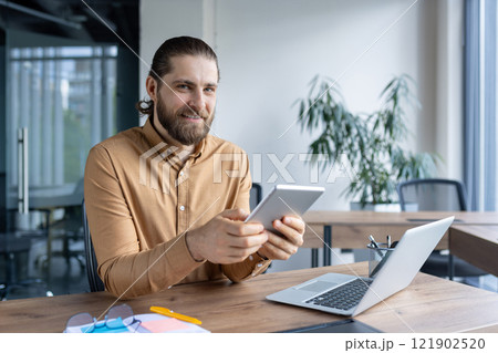 Smiling professional in casual attire working with a tablet in a modern office environment. A laptop is placed on the desk next to him, depicting a productive and professional setting. Smiling professional in casual attire working with a tablet in a modern office environment. A laptop is placed on the desk next to him, depicting a productive and professional setting. 121902520