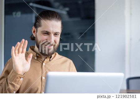 A bearded man in a casual shirt participates in a video call, waving and smiling warmly at the screen. Ideal representation for communication, remote work, or online professional interactions. 121902581