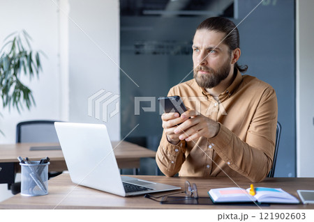 Focused man holds smartphone while working at an office desk with a laptop and documents. The office environment suggests analyzing, planning, and staying connected within a workplace. Focused man holds smartphone while working at an office desk with a laptop and documents. The office environment suggests analyzing, planning, and staying connected within a workplace. 121902603