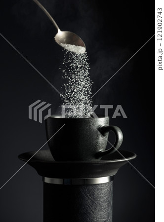 Sugar is poured into a black cup of coffee on a black background. 121902743