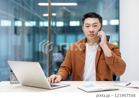 Portrait of a businessman inside the office at the workplace, a man is talking on the phone seriously and looking at the camera with concentration. Asian man at work with a laptop. Portrait of a businessman inside the office at the workplace, a man is talking on the phone seriously and looking at the camera with concentration. Asian man at work with a laptop. 121907588