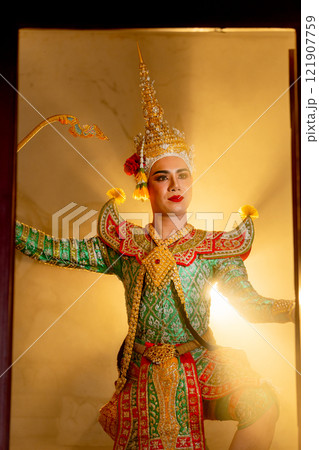 Vertical image of man with Khon mask charactor as deity stand with performance action in front of door frame of old building with beam warm light in the background. 121907759