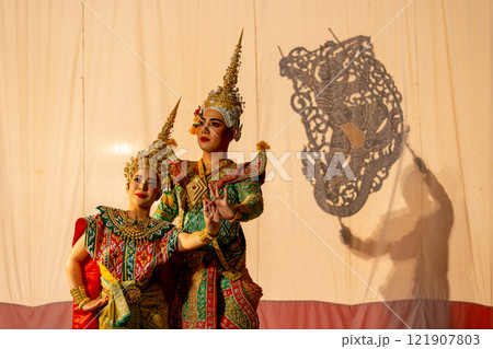 Man and woman with Khon or Mask dance costume stand with performance action in front of grand shadow play in the background with white screen. Man and woman with Khon or Mask dance costume stand with performance action in front of grand shadow play in the background with white screen. 121907803