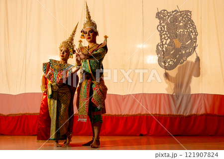 Two people of man and woman with Khon or Mask dance dress stand with performance action in front of grand shadow play in the background with white screen. 121907824