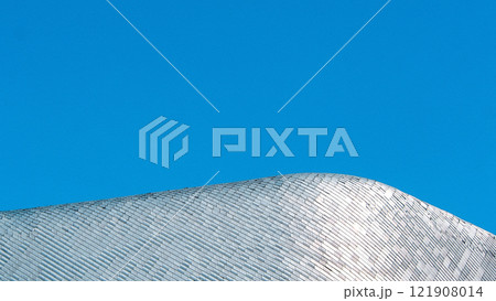 A sleek metallic roof with reflective tiles curves gracefully against a vivid blue sky, emphasizing minimalism and modern architecture 121908014