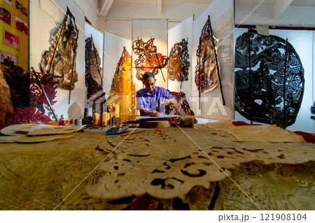 Wide shot of Asian senior man wear glasses use hammer and chisel to create grand shadow play craft and he concentrate to work in area of workplace with several products are place around workplace area 121908104