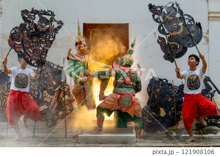 Two men with Khon mask charactor as angel and giant stand and performance action as figthing in front of door frame of old building also two men hold grand shadow play in front and warm light in back. 121908106