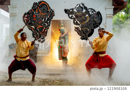 One man with Khon mask charactor as giant stand in front of door frame of old building also two men hold grand shadow play in front and warm light in back. 121908108