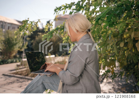 A woman is sitting on a bench with a laptop open in front of her. She is smiling and she is enjoying her time. Concept of relaxation and leisure. A woman is sitting on a bench with a laptop open in front of her. She is smiling and she is enjoying her time. Concept of relaxation and leisure. 121908168