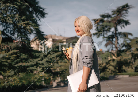 A woman is walking down the street with a white laptop bag and a cup of coffee. She is smiling and enjoying the sunny day. A woman is walking down the street with a white laptop bag and a cup of coffee. She is smiling and enjoying the sunny day. 121908170