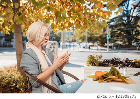 A blonde woman is sitting at a table with a cup of coffee and a plate of fruit. She is smiling and enjoying her time outside. 121908278