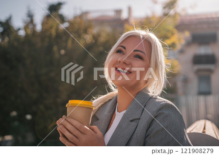 A blonde woman sits on a bench drinking coffee from a yellow cup. She is wearing a gray jacket and has her hair in a ponytail. The scene is peaceful and relaxing. 121908279