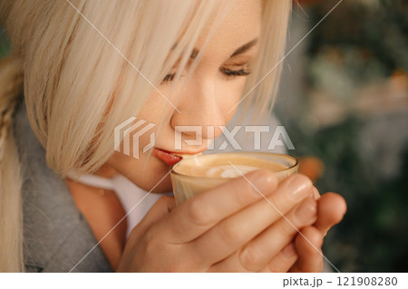 A blonde woman is drinking coffee from a glass. She is smiling and she is enjoying her drink. 121908280