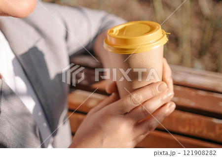 A person holding a coffee cup with a yellow lid. The person is wearing a gray jacket and white shirt. 121908282