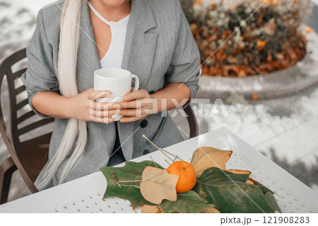 A woman is sitting at a table with a cup of coffee and a plate of oranges. The oranges are on a leaf, and the woman is wearing a gray jacket. Concept of relaxation and enjoyment. 121908283