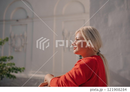A woman in a red sweater is sitting on a ledge. She is smiling and looking out at the street. A woman in a red sweater is sitting on a ledge. She is smiling and looking out at the street. 121908284