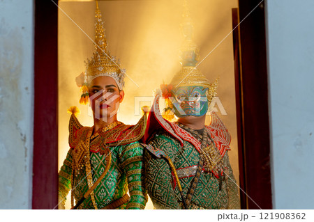 Two charactors of Khon mask costume stand with backs leaning against each other and human charactor stand in left side at door frame with beam light in the back. 121908362