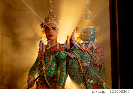 Wide shot of two charactors of Khon mask costume stand with backs leaning against each other and giant mask stand in right side at door frame with beam light in the back. Wide shot of two charactors of Khon mask costume stand with backs leaning against each other and giant mask stand in right side at door frame with beam light in the back. 121908363