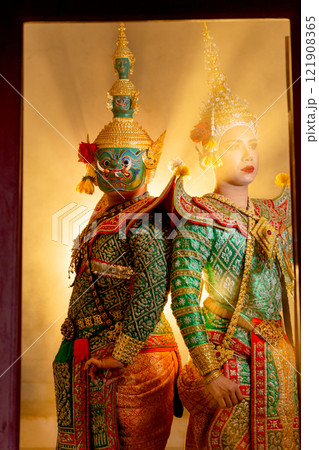Vertical image of two charactors of Khon mask costume stand with backs leaning against each other at door frame with beam light in the back. 121908365