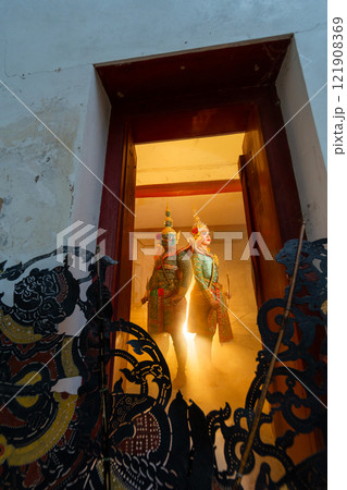 Vertical image and wide shot of two charactor of Khon mask costume stand with backs leaning against each other at door frame with light in the back and grand shadow play crafts in the front. Vertical image and wide shot of two charactor of Khon mask costume stand with backs leaning against each other at door frame with light in the back and grand shadow play crafts in the front. 121908369