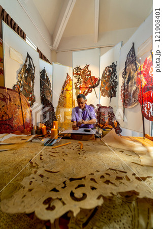 Vertical image Asian senior man wear glasses use hammer and chisel to create grand shadow play craft and he concentrate to work in area of workplace with several products are place around workplace. 121908401