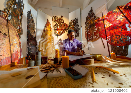 Side view of Asian senior man wear glasses use hammer and chisel to create grand shadow play craft and he concentrate to work in area of workplace with several products are place around workplace area 121908402