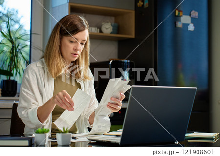 Stressed woman reviewing bills at home. Female organizing receipts, sitting at kitchen table with laptop and paper documents. Concept of budgeting and managing personal finances 121908601
